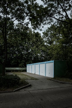 A row of five white garage doors with blue frames aligned in a line, set in a wooded environment. The garages are situated on a paved area surrounded by dense greenery, including tall trees with rich foliage. The sky above is overcast, casting a moody ambiance across the scene.