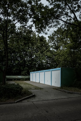 A row of five white garage doors with blue frames aligned in a line, set in a wooded environment. The garages are situated on a paved area surrounded by dense greenery, including tall trees with rich foliage. The sky above is overcast, casting a moody ambiance across the scene.