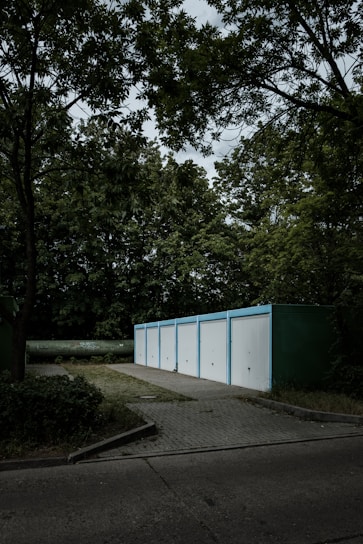 A row of five white garage doors with blue frames aligned in a line, set in a wooded environment. The garages are situated on a paved area surrounded by dense greenery, including tall trees with rich foliage. The sky above is overcast, casting a moody ambiance across the scene.