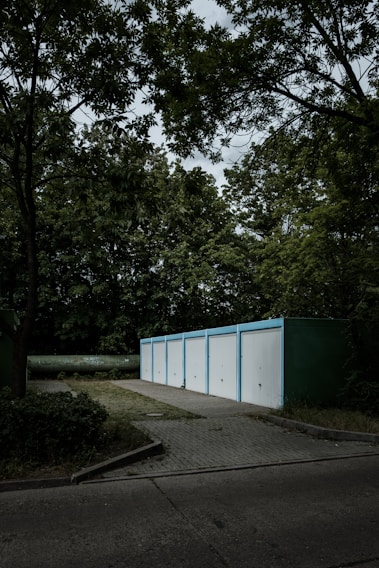 A row of five white garage doors with blue frames aligned in a line, set in a wooded environment. The garages are situated on a paved area surrounded by dense greenery, including tall trees with rich foliage. The sky above is overcast, casting a moody ambiance across the scene.