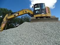 A large excavator stands atop a pile of gravel, with a clear blue sky and some trees in the background. The machine appears to be actively engaged in a construction or excavation task.