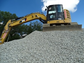 A large excavator stands atop a pile of gravel, with a clear blue sky and some trees in the background. The machine appears to be actively engaged in a construction or excavation task.