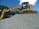 A large excavator stands atop a pile of gravel, with a clear blue sky and some trees in the background. The machine appears to be actively engaged in a construction or excavation task.