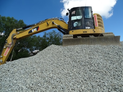 A large excavator stands atop a pile of gravel, with a clear blue sky and some trees in the background. The machine appears to be actively engaged in a construction or excavation task.