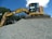 A large excavator stands atop a pile of gravel, with a clear blue sky and some trees in the background. The machine appears to be actively engaged in a construction or excavation task.