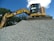 A large excavator stands atop a pile of gravel, with a clear blue sky and some trees in the background. The machine appears to be actively engaged in a construction or excavation task.