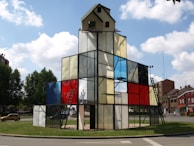 A geometric glass structure resembling a building, with a small house-like top and metal ladders. Panels of stained glass in various colors, including red, blue, and clear, are used for walls. The structure is situated in a roundabout surrounded by greenery, with a few residential buildings in the background under a partly cloudy sky.