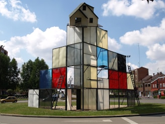 A geometric glass structure resembling a building, with a small house-like top and metal ladders. Panels of stained glass in various colors, including red, blue, and clear, are used for walls. The structure is situated in a roundabout surrounded by greenery, with a few residential buildings in the background under a partly cloudy sky.