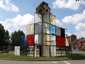 A geometric glass structure resembling a building, with a small house-like top and metal ladders. Panels of stained glass in various colors, including red, blue, and clear, are used for walls. The structure is situated in a roundabout surrounded by greenery, with a few residential buildings in the background under a partly cloudy sky.