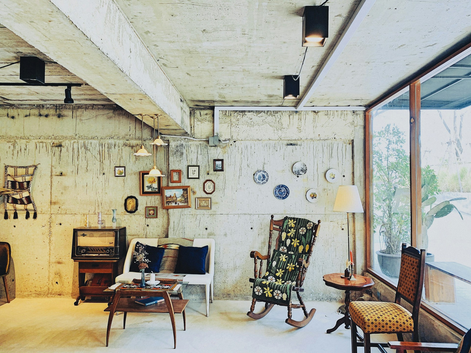 A cozy and eclectic interior featuring a vintage style setting. The room has a rustic concrete wall adorned with a variety of small framed artworks and decorative plates. A mix of furniture includes a floral-patterned rocking chair, a small white loveseat with dark cushions, and a wooden side table with a retro radio on it. Hanging pendant lights illuminate the space, and a large window lets in natural light, offering a view of greenery outside. The decor includes a wall tapestry and various knick-knacks providing a warm, nostalgic atmosphere.