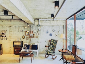 A cozy and eclectic interior featuring a vintage style setting. The room has a rustic concrete wall adorned with a variety of small framed artworks and decorative plates. A mix of furniture includes a floral-patterned rocking chair, a small white loveseat with dark cushions, and a wooden side table with a retro radio on it. Hanging pendant lights illuminate the space, and a large window lets in natural light, offering a view of greenery outside. The decor includes a wall tapestry and various knick-knacks providing a warm, nostalgic atmosphere.