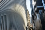A modern architectural scene featuring a large building with a distinct, flowing design composed of white vertical slats. Sunlight creates patterns of light and shadow on the structure. In the foreground, modern glass and steel buildings reflect the light. Skyscrapers are visible in the background, hinting at an urban environment.