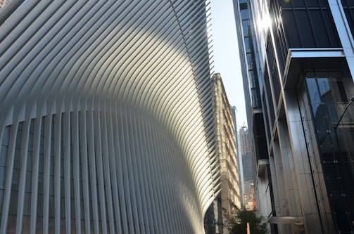 A modern architectural scene featuring a large building with a distinct, flowing design composed of white vertical slats. Sunlight creates patterns of light and shadow on the structure. In the foreground, modern glass and steel buildings reflect the light. Skyscrapers are visible in the background, hinting at an urban environment.