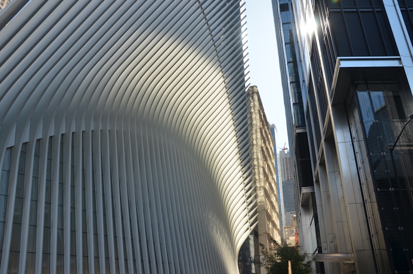 A modern architectural scene featuring a large building with a distinct, flowing design composed of white vertical slats. Sunlight creates patterns of light and shadow on the structure. In the foreground, modern glass and steel buildings reflect the light. Skyscrapers are visible in the background, hinting at an urban environment.