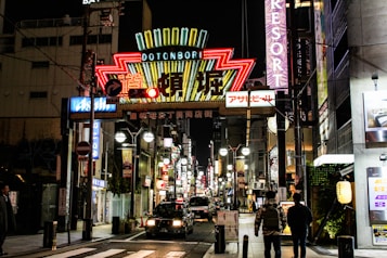 A vibrant city street scene at night with a large, colorful neon sign displaying the word 'Dotonbori' in bright lights. The street is bustling with activity, featuring numerous illuminated advertisements and storefronts. People are walking along the sidewalk, and a car is driving down the street. The atmosphere is lively and bustling, typical of an urban nightlife setting.