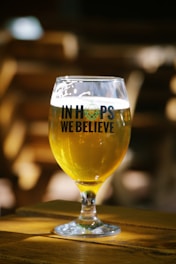 A glass of beer sits on a wooden table, filled with a golden liquid. The glass features the words 'In Hops We Believe' in bold, dark lettering. Natural light highlights the beer's vibrant color, creating a warm, inviting atmosphere. The background is intentionally blurred, directing focus to the glass.