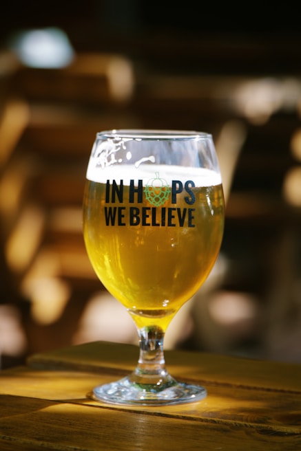 A glass of beer sits on a wooden table, filled with a golden liquid. The glass features the words 'In Hops We Believe' in bold, dark lettering. Natural light highlights the beer's vibrant color, creating a warm, inviting atmosphere. The background is intentionally blurred, directing focus to the glass.