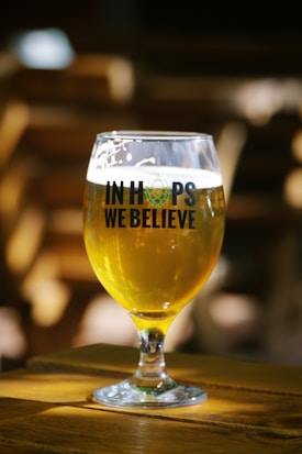 A glass of beer sits on a wooden table, filled with a golden liquid. The glass features the words 'In Hops We Believe' in bold, dark lettering. Natural light highlights the beer's vibrant color, creating a warm, inviting atmosphere. The background is intentionally blurred, directing focus to the glass.