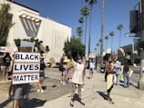 A group of people are gathered on a sunny day, holding signs with messages supporting the Black Lives Matter movement. They stand on a street corner in front of a large building and palm trees. Some signs read 'Black Lives Matter' and 'End Police Brutality'. The individuals are wearing masks and casual clothing.