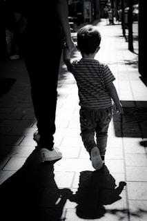A silhouette of an adult and a child holding hands while walking down a sunlit sidewalk. The child wears striped clothing, and the adult is dressed in dark pants with white sneakers. The scene is captured in black and white, with long shadows casting ahead of them.