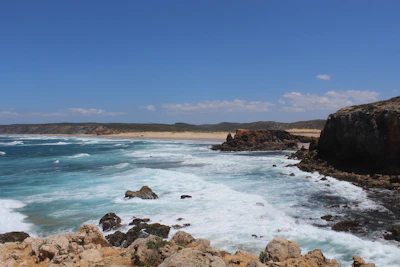 A coastal scene featuring rugged cliffs meeting turquoise waters, with waves crashing against the shore under a teal-tinted sky.