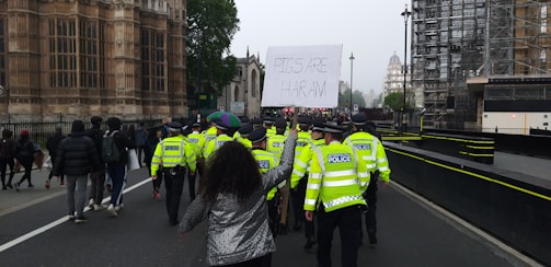 A group of police officers in high-visibility jackets are leading a procession of people on a street next to a historic building. One person in the crowd is holding a large sign that reads 'PIGS ARE HARAM'. The sky appears to be overcast, and the atmosphere seems tense.