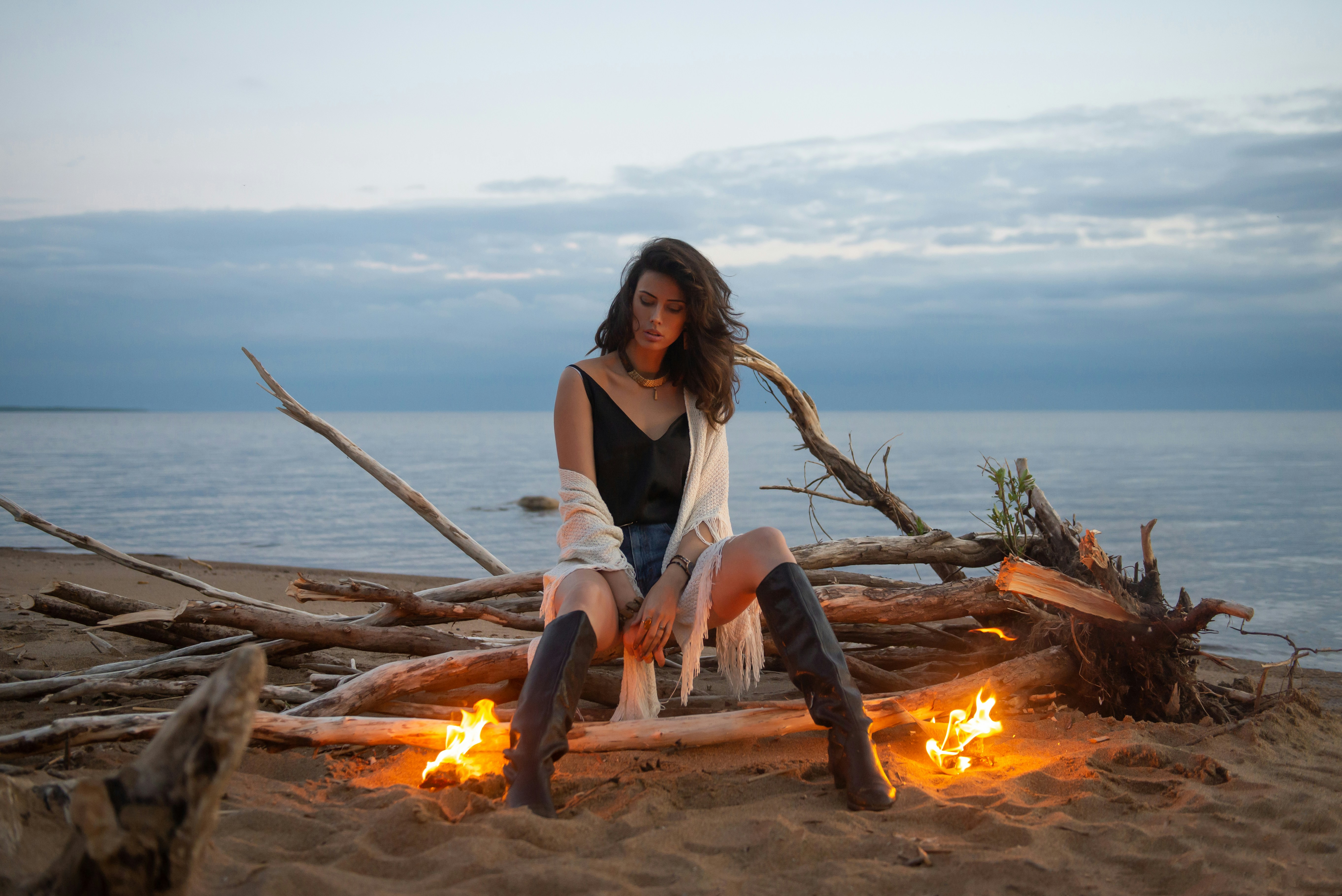 woman in white tank top sitting on brown log near bonfire during daytime flame teams background