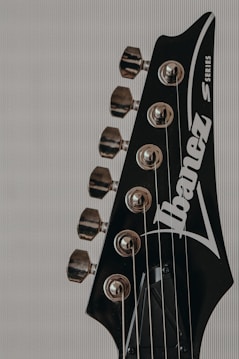 The image features a close-up view of the headstock of an electric guitar. The headstock is black with chrome tuning pegs and string winds visible. The brand name is prominently displayed in white lettering across the surface. The strings extend down from the tuning pegs.