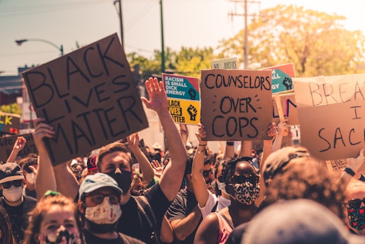 A large group of people gathered outdoors, holding various signs with messages advocating for social justice. Some signs include phrases like 'Black Lives Matter' and 'Counselors Over Cops.' Many participants are wearing masks, reflecting a sense of unity in protest.