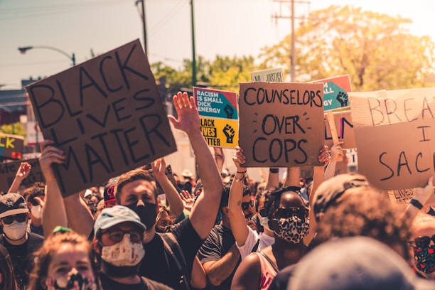 A large group of people gathered outdoors, holding various signs with messages advocating for social justice. Some signs include phrases like 'Black Lives Matter' and 'Counselors Over Cops.' Many participants are wearing masks, reflecting a sense of unity in protest.