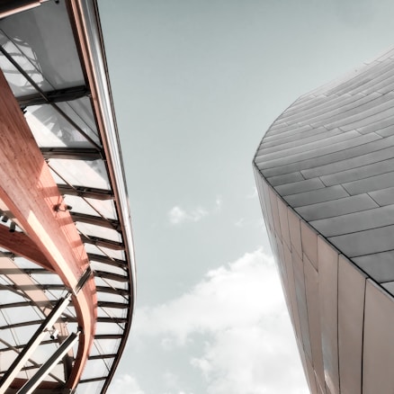Two modern architectural structures with curved designs rise against a pale sky. The structure on the left features a combination of wood and glass with visible steel supports, creating an open appearance. The building on the right displays metallic panels with a sleek, futuristic design.