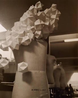 A mannequin torso with a complex geometric structure attached to it, composed of interconnected polygonal shapes. The background appears to show another mannequin and a workspace with dim lighting.