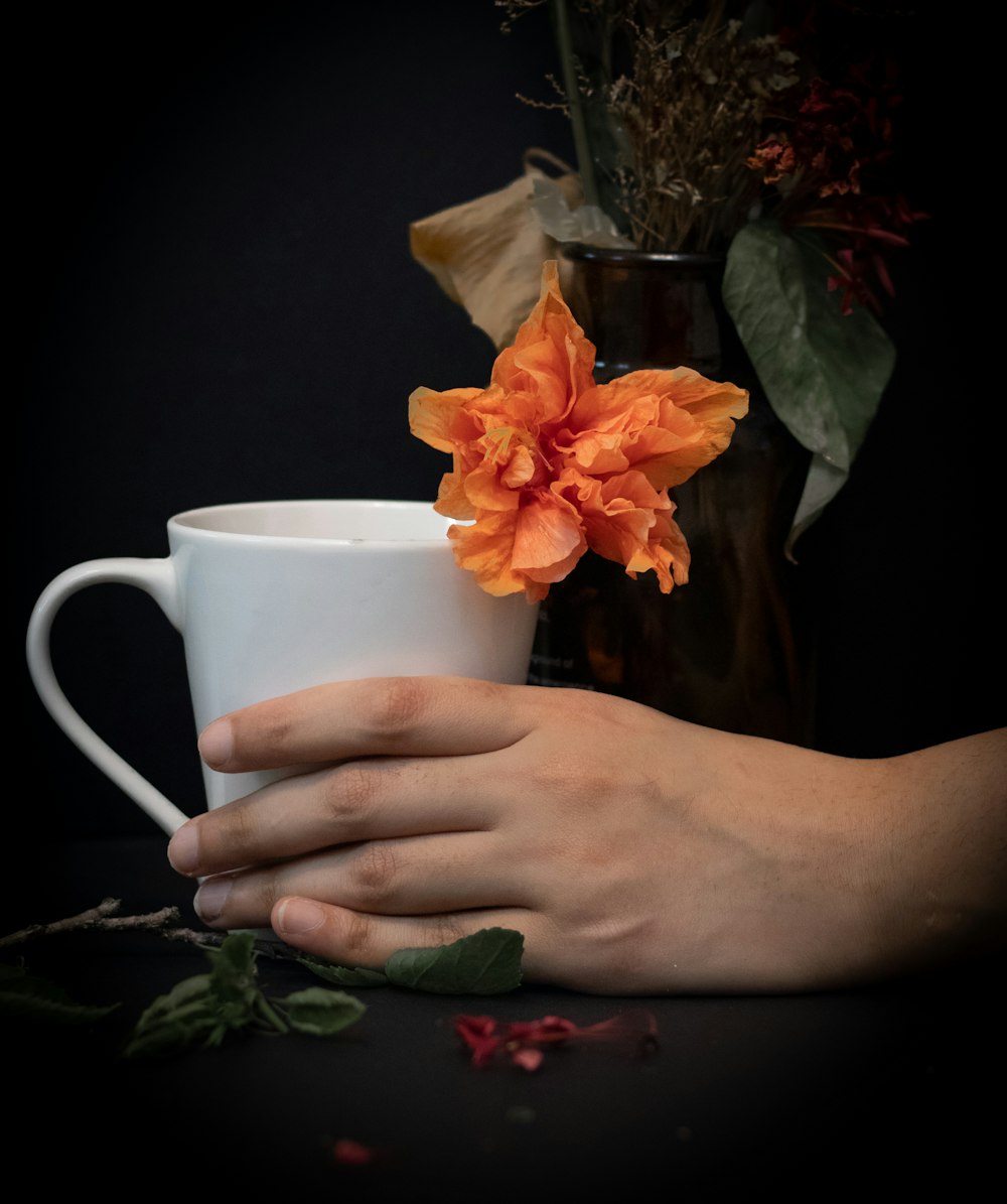 person holding white ceramic mug with orange flower