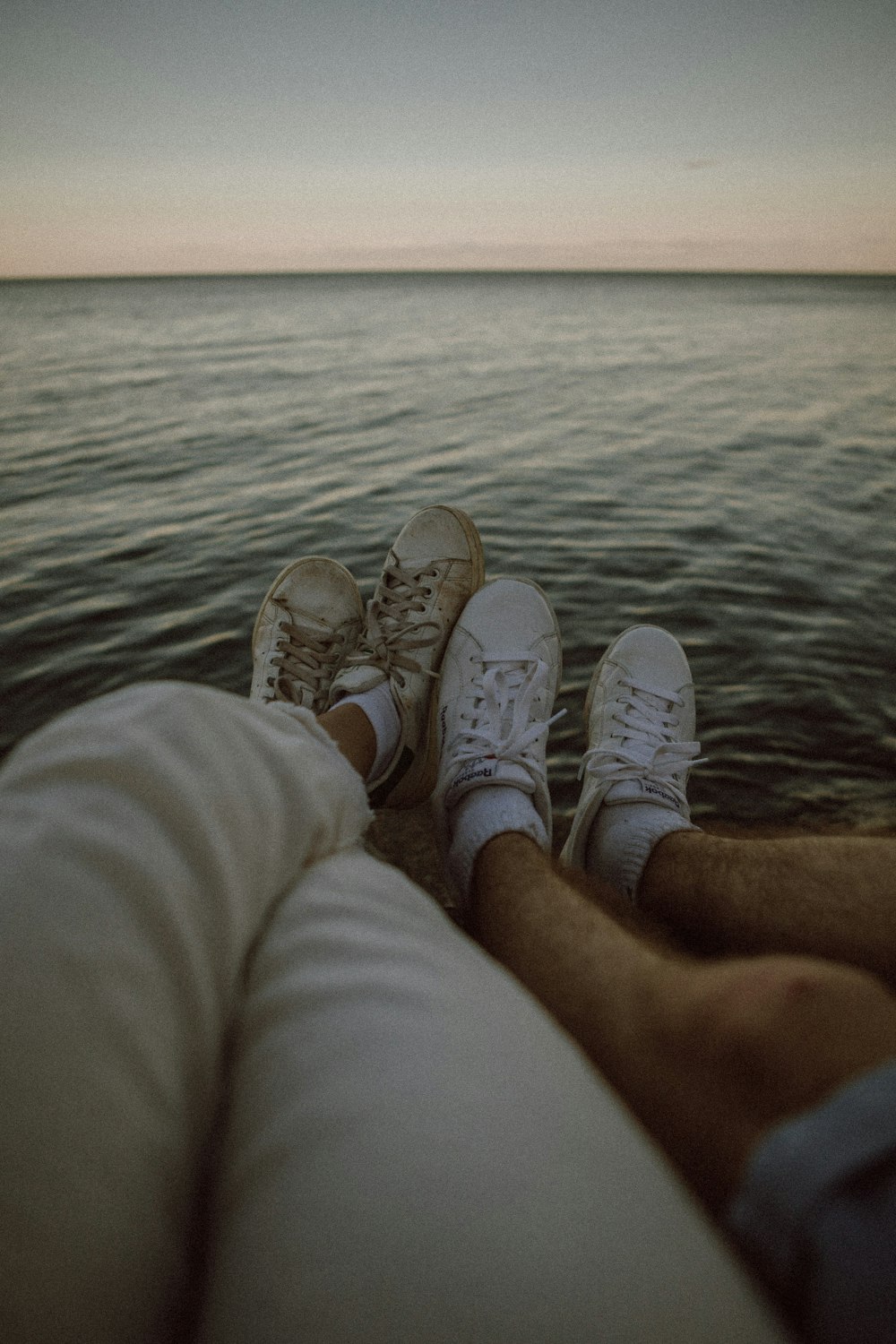 person in white pants and white sneakers sitting on rock near body of water during daytime