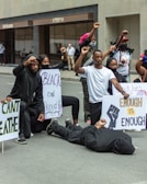 A group of people are participating in a protest, holding signs with messages related to social justice. One person is lying on the ground as others kneel or raise fists in solidarity.
