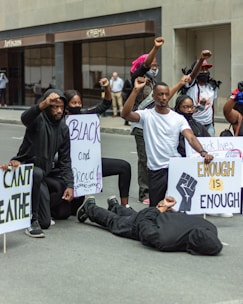 A group of people are participating in a protest, holding signs with messages related to social justice. One person is lying on the ground as others kneel or raise fists in solidarity.