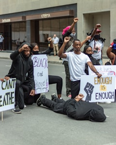 A group of people are participating in a protest, holding signs with messages related to social justice. One person is lying on the ground as others kneel or raise fists in solidarity.