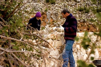 Two individuals are engaged in outdoor work amidst a rocky landscape. One person, wearing a purple hat, appears to be bending over while another stands nearby, wearing gloves and observing or handling the rocks. The setting is natural, with leafy branches in the foreground and a backdrop of stone and sparse vegetation.