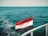 A flag with red, white, and black horizontal stripes featuring a golden emblem is prominently displayed on a boat, with the flag waving in a breeze over a vast expanse of deep blue ocean under a clear sky.