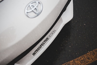 A close-up of a car with the Toyota logo on the hood. The car appears to be white with an inscription 'INTROSPECTIVEDESIGN' near the front bumper. The background shows an asphalt road with a yellow line.