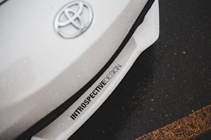 A close-up of a car with the Toyota logo on the hood. The car appears to be white with an inscription 'INTROSPECTIVEDESIGN' near the front bumper. The background shows an asphalt road with a yellow line.