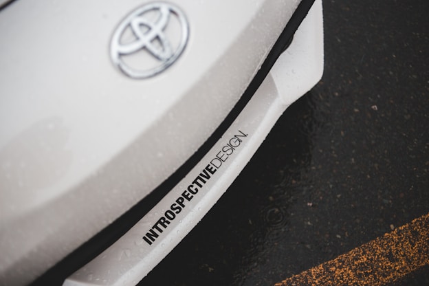 A close-up of a car with the Toyota logo on the hood. The car appears to be white with an inscription 'INTROSPECTIVEDESIGN' near the front bumper. The background shows an asphalt road with a yellow line.