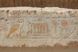 Carved stone relief with ancient Egyptian hieroglyphics, including an owl, a seated figure, a sun disk, and other symbols.