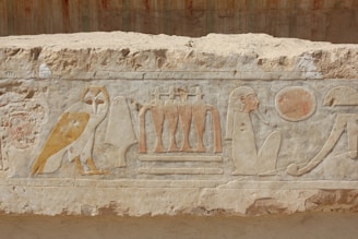 Carved stone relief with ancient Egyptian hieroglyphics, including an owl, a seated figure, a sun disk, and other symbols.