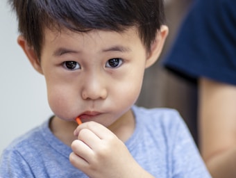 A young child with short dark hair is looking directly at the camera while holding a small orange object to their mouth. The child is wearing a blue shirt and appears to be in an indoor setting.