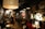 A dimly lit café with a cozy atmosphere, featuring several groups of people sitting at small tables. Large pendant lights hang from the ceiling, casting a warm glow. The décor includes a world map on the wall, various plants, and shelves filled with books and decorative items. The seating arrangement includes both couches and chairs, providing a comfortable setting for conversation and relaxation.