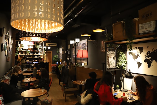 A dimly lit café with a cozy atmosphere, featuring several groups of people sitting at small tables. Large pendant lights hang from the ceiling, casting a warm glow. The décor includes a world map on the wall, various plants, and shelves filled with books and decorative items. The seating arrangement includes both couches and chairs, providing a comfortable setting for conversation and relaxation.