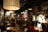 A dimly lit café with a cozy atmosphere, featuring several groups of people sitting at small tables. Large pendant lights hang from the ceiling, casting a warm glow. The décor includes a world map on the wall, various plants, and shelves filled with books and decorative items. The seating arrangement includes both couches and chairs, providing a comfortable setting for conversation and relaxation.