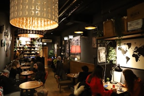 A dimly lit café with a cozy atmosphere, featuring several groups of people sitting at small tables. Large pendant lights hang from the ceiling, casting a warm glow. The décor includes a world map on the wall, various plants, and shelves filled with books and decorative items. The seating arrangement includes both couches and chairs, providing a comfortable setting for conversation and relaxation.