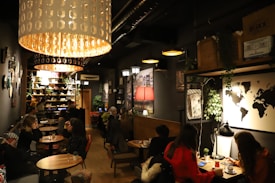 A dimly lit caf&eacute; with a cozy atmosphere, featuring several groups of people sitting at small tables. Large pendant lights hang from the ceiling, casting a warm glow. The d&eacute;cor includes a world map on the wall, various plants, and shelves filled with books and decorative items. The seating arrangement includes both couches and chairs, providing a comfortable setting for conversation and relaxation.